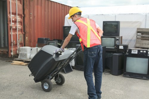 A man wearing a yellow safety helmet, a high-visibility vest with red and yellow stripes, a white t-shirt, and blue jeans is pushing a black wheeled waste bin on a trolley at an outdoor storage or disposal site. Behind him, there are various discarded electronic appliances, including old televisions, computer monitors, and stereo systems, stacked against a red shipping container and wooden pallets. The area appears to be a designated rubbish removal or recycling zone, possibly part of an office clearance service in Carshalton, with a mix of outdoor industrial surroundings and waste materials set on a paved surface under natural daylight.