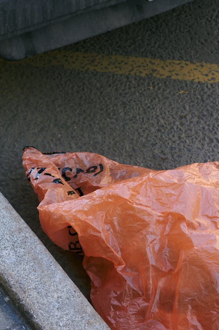 An orange plastic rubbish bag with black text partially visible lies crumpled on a dark, textured asphalt pavement, close to a grey concrete curb. The bag appears to be filled with waste and is restin